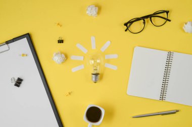 Düz boş klip Board, ofis malzemeleri, kalem, Not defteri, Gözlük, kahve fincan, sarı arka planda ampul ile dağınık masa üst görüntü beyin fırtınası mali planlama yatıyordu.