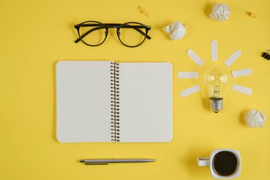 Boş klip panosu, ofis malzemeleri, kalem, Not defteri, Gözlük, kahve bardağı ve sarı arka planda ampul ile Office çalışma alanı. Konsept beyin fırtınası ve yeni fikir