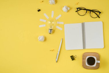 Düz boş klip Board, ofis malzemeleri, kalem, Not defteri, Gözlük, kahve fincan, sarı arka planda ampul ile dağınık masa üst görüntü beyin fırtınası mali planlama yatıyordu.