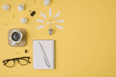 Düz boş klip Board, ofis malzemeleri, kalem, Not defteri, Gözlük, kahve fincan, sarı arka planda ampul ile dağınık masa üst görüntü beyin fırtınası mali planlama yatıyordu.