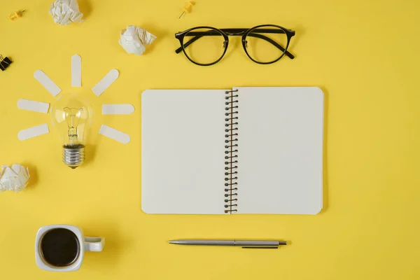 Office workspace with blank clip board, office supplies, pen, notepad, eyeglasses, coffee cup and light bulb on yellow background. Concept brainstorming and new idea