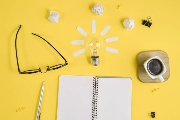 Office workspace with blank clip board, office supplies, pen, notepad, eyeglasses, coffee cup and light bulb on yellow background. Concept brainstorming and new idea