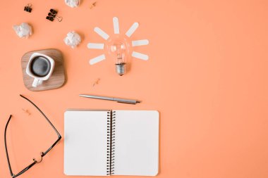 Düz boş klip Board, ofis malzemeleri, kalem, Not defteri, Gözlük, kahve fincan, turuncu arka planda ampul ile dağınık masa üst görüntü beyin fırtınası mali planlama yatıyordu.