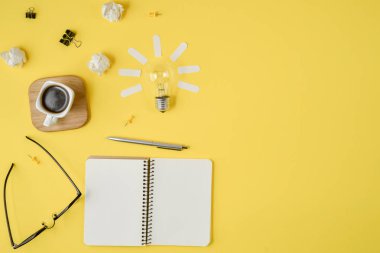 Düz boş klip Board, ofis malzemeleri, kalem, Not defteri, Gözlük, kahve fincan, sarı arka planda ampul ile dağınık masa üst görüntü beyin fırtınası mali planlama yatıyordu.