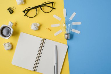 Düz boş klip Board, ofis malzemeleri, kalem, Not defteri, Gözlük, kahve bardağı, sarı ve mavi arka planda ampul ile dağınık masa üst görüntü beyin fırtınası mali planlama yatıyordu.
