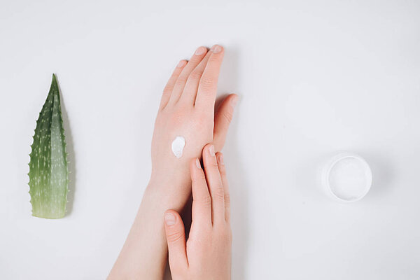 Closeup of beautiful female hands applying cream. Organic cream from Aloe vera fresh leaves on white background. Flat lay, top view, copy space. Healthcare concept.