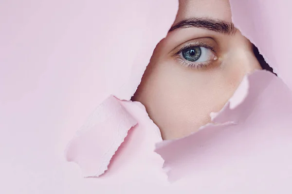 Primer plano de un ojo femenino que mirando se ve a través de un agujero en un papel rosa. Cosmética profesional y cuidado facial. Fondo de color brillante y un hueco en el papel —  Fotos de Stock