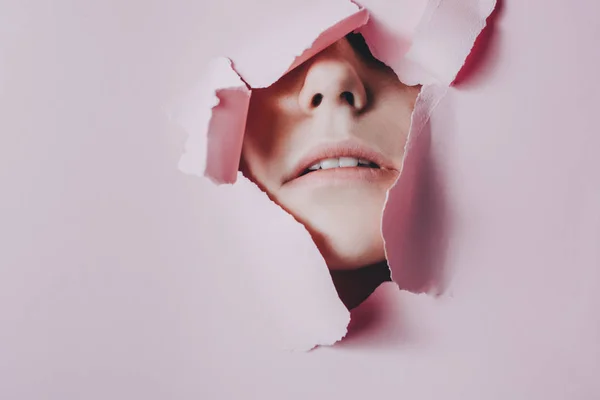 Imágenes creativas de bellos labios femeninos que se pueden ver a través de un agujero en papel rosa. Cosmética profesional y cuidado facial. Fondo de color brillante y un hueco en el papel —  Fotos de Stock