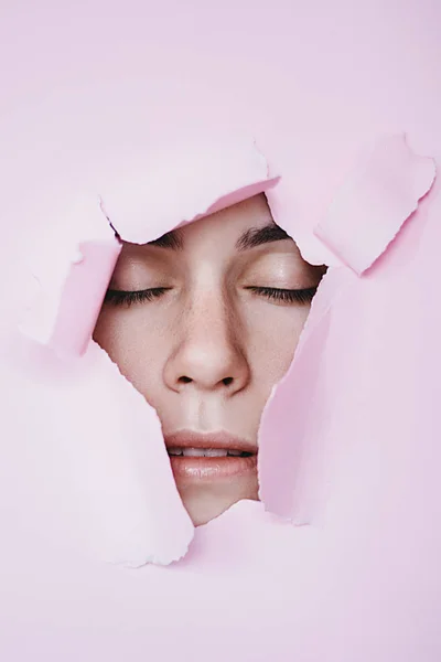 Imagen creativa de una joven hermosa con los ojos cerrados miradas en un agujero en papel rosa. Cosmética profesional y cuidado facial. Fondo de color brillante y un hueco en el papel — Foto de Stock