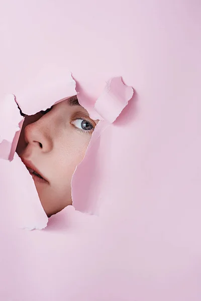 La cara de la muchacha joven hermosa mira a través del agujero en el papel rosado. Cosmética profesional y cuidado facial. Fondo de color brillante y un hueco en el papel. Espacio para texto —  Fotos de Stock