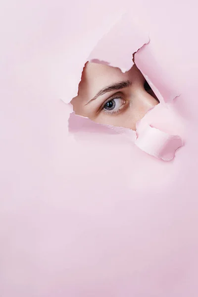 Close up of a female eye that looking is seen through a hole in torn pink paper. Beauty salon advertising with copy space. Creative and fashion concept.