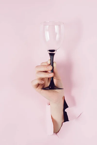 La mano femenina sostiene la copa de vino se ve a través de un agujero en papel rosa roto. Vista aérea, espacio de copia. Concepto de vacaciones y cumpleaños . — Foto de Stock