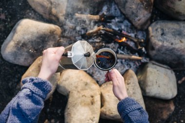 Genç kadının yatay açık hava görüntüsü şenlik ateşine yakın sıcak içecek döker. Genç bir turist kadın bir fincan sıcak bir içecek içecekler ve tatil de doğanın tadını çıkarıyor. Kamp konsepti