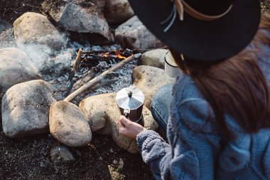 Şapkalı gezgin kız şenlik ateşine yakın bir ormanda sıcak kahve tutar. Kadın otururve yürüyüşten sonra bir fincan kahve tutar. Trekking konsepti