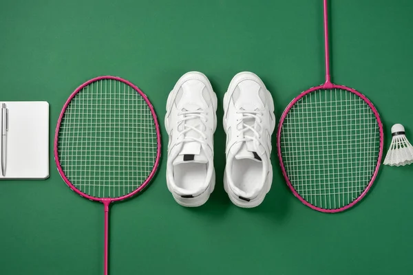 Sports flat lay with shuttlecock and badminton racket, sneakers, pen and notepad on green background. Fitness, sport and healthy lifestyle concept.