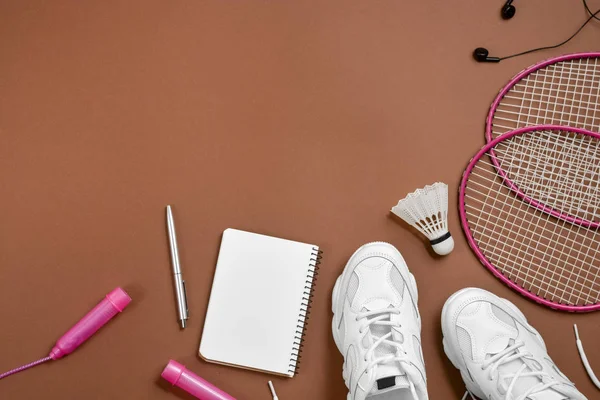 Deportivo plano con transbordador y raqueta de bádminton, saltar la cuerda, zapatillas de deporte, auriculares, bolígrafo y bloc de notas sobre fondo marrón. Fitness, deporte y estilo de vida saludable . — Foto de Stock