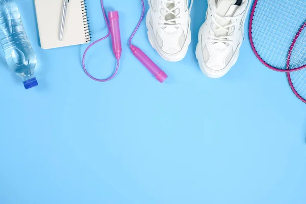 Sports flat lay with badminton racket, skipping rope, sneakers, bottle water, pen and notepad on blue background. Fitness, sport and healthy lifestyle concept.