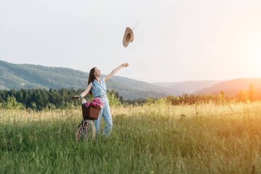 Bir şapka atma genç mutlu gülümseyen kadın Açık portre ve gün batımı sırasında dağ doğa sahiptir. Elinde çiçek sepeti olan bir bisiklet tutuyor. Konsept açık doğa yaşam tarzı