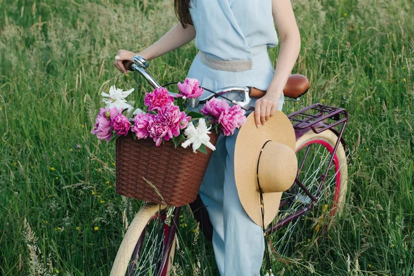 Outdoor horizontal image of female beautiful hands hold a bicycle with a basket of red and white flowers and a stylish hat on the background of nature. Concept outdoor nature lifestyle.