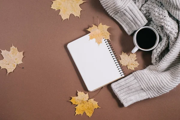 Werkruimte met gouden esdoorn bladeren, notebook, koffie kopje, trui op bruine achtergrond. Creatieve compositie. Herfst-of winter concept. Platte lay, bovenaanzicht — Stockfoto