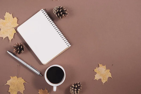 Herfst samenstelling. Gouden esdoorn bladeren, koffie kopje, hobbels, notebook en pen op bruine achtergrond. Herfst-of winter concept. Platte lay, bovenaanzicht — Stockfoto