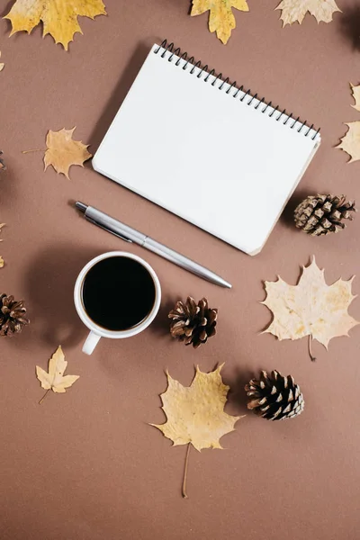 Werkruimte met gouden esdoorn bladeren, koffiekop, hobbels, notebook en pen op bruine achtergrond. Creatieve compositie. Herfst-of winter concept. Platte lay, bovenaanzicht — Stockfoto