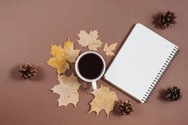 Herfst samenstelling. Kopje koffie, notebook, herfst esdoorn bladeren en hobbels op bruine achtergrond. Platte lay, bovenaanzicht, kopieer ruimte. — Stockfoto