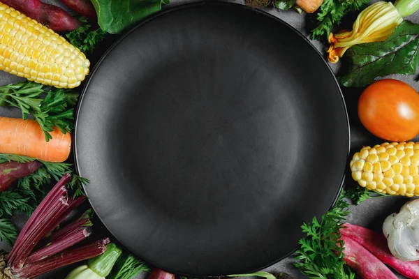 Assorted fresh vegetables on black background. Corn, tomatoes, zucchini, carrot, beetroot, garlic, bean, black plate. Health lifestyle and food concept. Flat lay, top view, copy space