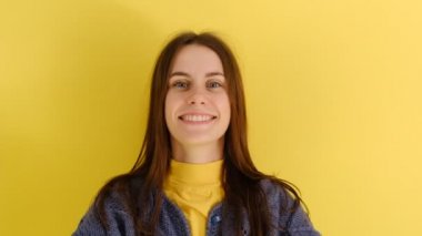 Mavi kazak giyen mutlu bir kadın şapkasını giyer, mutlu bir şekilde gülümser, sarı arka planda izole bir şekilde. Beyaz kadın içeride poz veriyor.