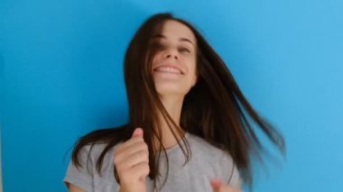 Uçan saçı geniş bir şekilde gülümseyen, el çırparak kameraya bakan, mavi arka planda poz veren genç ve güzel bir kızın yakın çekimi. İnsanlar samimi duygular yaşam tarzı.