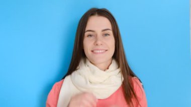 Mavi arka planda soyutlanmış, pembe kazak giyen ve eşarp takan mutlu genç bir kadın. Kameraya bak.