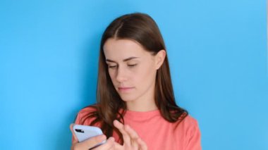 Cep telefonunda mesaj yazan neşeli bir kız, çevrimiçi iletişimden hoşlanıyor, geri bildirim yapıyor, pembe kazak giyiyor, mavi stüdyo duvarında izole edilmiş. Teknoloji konsepti