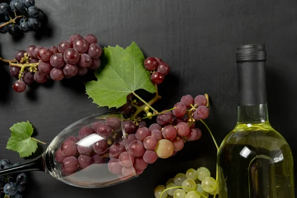 A bottle, wine glasses, fresh grapes and leaves on black background. Flat lay, top view, copy space. Wine bar, winery, wine tasting concept