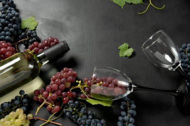 Beyaz şarap şişesi, iki şarap kadehi, taze üzüm ve siyah ahşap arka plan üzerinde yaprakları. Düz döşeme, üst görünüm, kopyalama alanı. Şarap barı, şaraphane, şarap tatma konsepti