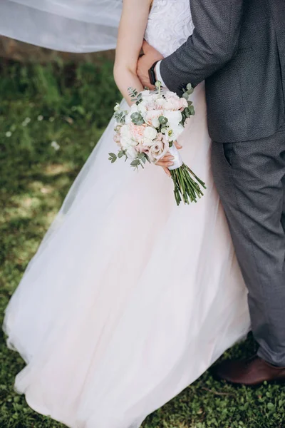 Közelről menyasszony egy fehér ruhában, és a vőlegény egy öltönyt ölelgetés, és tart egy szép csokor fehér, rózsaszín virágok és zöld, díszített. Esküvői részletek és természetes koncepció. — Stock Fotó