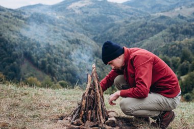 Kırmızı ceketli genç adam, örgü bere brunch hazırlamak, dağlarda şenlik ateşi için. Sakallı gezgin adam, kamp ateşinin yanında oturuyor. Seyahat gerçek vahşi yaşam tarzı keşfedin