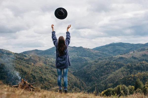 Η πίσω όψη της νεαρής γυναίκας περπατά σε όλο το χωράφι και πετάει το καπέλο της στο φόντο της οροσειράς του βουνού, ανεβάζει τα χέρια στον αέρα, τη νεολαία και την ευτυχία. Εξερευνήστε τον πραγματικό τρόπο ζωής της άγριας φύσης — Φωτογραφία Αρχείου