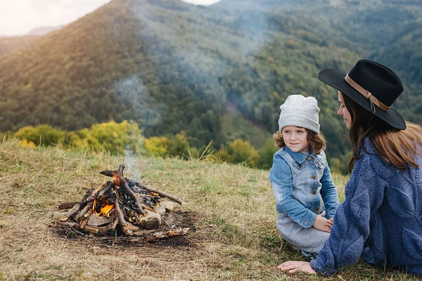 Οριζόντια εξωτερική εικόνα της νεαρής μητέρας και της ομορφης κόρης της που κάθονται κοντά στη φωτιά στα βουνά. Ταξίδια, άνθρωποι και υγιής τρόπος ζωής. Καυκάσια γυναίκα με μωρό στα βουνά — Φωτογραφία Αρχείου