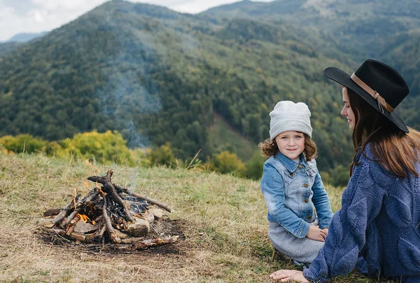 Μια νεαρή μητέρα και η όμορφη ευτυχισμένη κόρη της που κάθονταν κοντά στη φωτιά στα βουνά. Ταξίδια, άνθρωποι και υγιής τρόπος ζωής. Καυκάσια γυναίκα με μωρό στα βουνά — Φωτογραφία Αρχείου