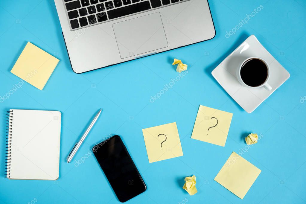 Question mark on sticky notes, notebook, laptop, cup coffee, phone, pen on blue background. FAQ concept. Working with clients. Troubles in work. Flat lay, top view, copy space