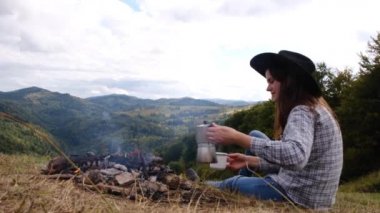 Kamp ateşinin yanında oturan tatmin olmuş hippi bayan gezgin, dağların manzarası, güzel ve ilham verici kamp yeri ile kahve makinesinden kahve doldurur. Sakin