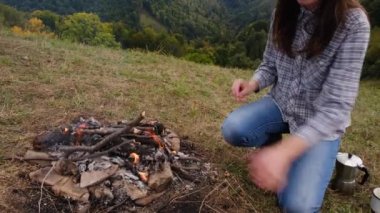 Güzel genç bir kadın, dağlarda kahve yaptığı için kamp ateşini yakıyor. Kamp ateşinin yanında oturan gezgin kız. Gerçek vahşi yaşam tarzını keşfet.