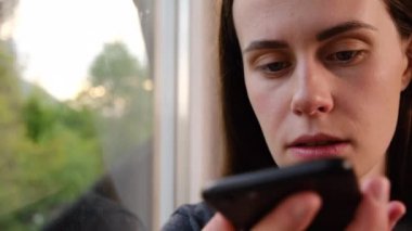 Ciddi esmer kadın elinde telefonla pencere eşiğinde oturuyor. Sanal dijital ses asistanını aktifleştiriyor, sesli mesaj kaydediyor ya da akıllı telefondan hoparlörden konuşuyor. Teknoloji konsepti