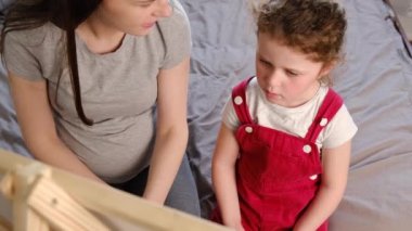 Hamile, genç bir annenin yan görüntüsü güzel bir anaokulu kızına evde yatakta otururken tahtaya yazmayı ve aile eğitimini öğretiyor. Erken çocukluk gelişimi.