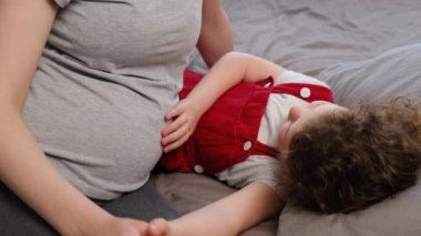 Sevimli, gülümseyen küçük bir kız ellerini okşuyor ve bebeğin annesinin karnını tekmelemesini dinliyor. Sevgi dolu bir anne ve güzel bir anaokulu kızı evde rahat bir yatakta oturup birlikte vakit geçiriyorlar..