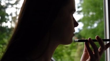Pencere pervazında oturan genç bir kadının yan görüntüsü telefonda sanal dijital ses asistanını aktifleştir, kız arkadaşlarıyla online veya hoparlörde sohbet et, sesli mesaj kaydet