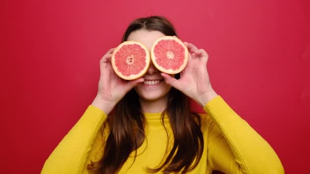 照片中 年轻的微笑的女人把一半柚子放在她的眼睛里 滑稽的漂亮姑娘 长着柑橘类新鲜的一半水果 穿着黄色的毛衣 在红色的工作室背景下被隔离 — 图库视频影像