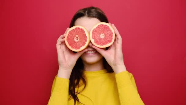 喜びに満ちた若い女性は 柑橘系の果物の2つのスライスを保持し 新鮮なグレープフルーツの匂いを楽しんで かわいい陽気な女の子はカメラを見て笑って セーターを着て 赤い壁の背景に隔離されたポーズ — ストック動画