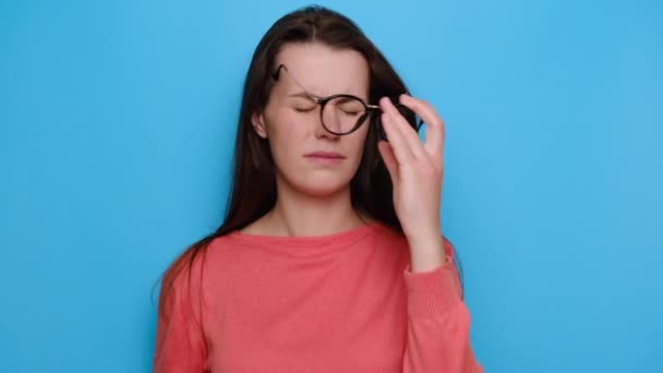 厌倦了眼镜的年轻女子按摩鼻桥 疲倦的女士头痛或偏头痛 感觉眼力紧张 穿着红色毛衣 与蓝色背景隔离 眼睛疲劳概念 — 图库视频影像