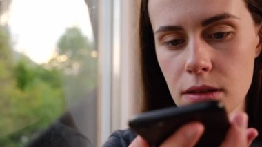 Ciddi esmer kadın elinde telefonla pencere eşiğinde oturuyor. Sanal dijital ses asistanını aktifleştiriyor, sesli mesaj kaydediyor ya da akıllı telefondan hoparlörden konuşuyor. Teknoloji konsepti